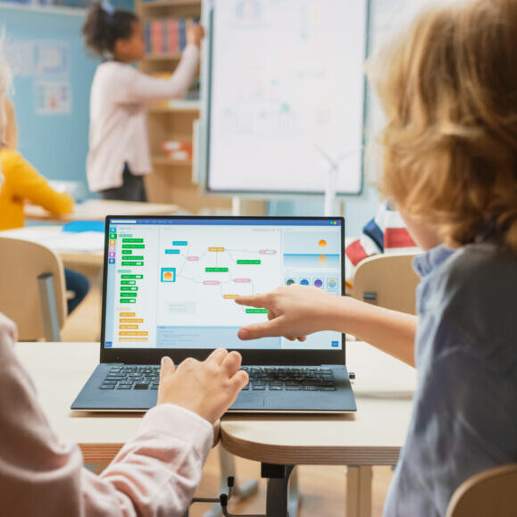 Elementary School Science Class: Over the Shoulder Little Boy and Girl Use Laptop with Screen Showing Programming Software. Physics Teacher Explains Lesson to a Diverse Class full of Smart Kids